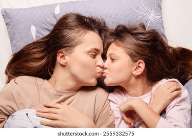 A mother and her young daughter lie closely together in bed, smiling and playfully making kissy faces. Sunlight filters softly into the room, creating a warm and loving atmosphere. - Powered by Shutterstock