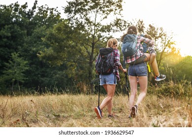 Mother Hiking With Her Two Daughter.They Walking Over The Meadows And Joying In Sunset.Older Sister Carrying His Younger Sister On Her Back.rear View.