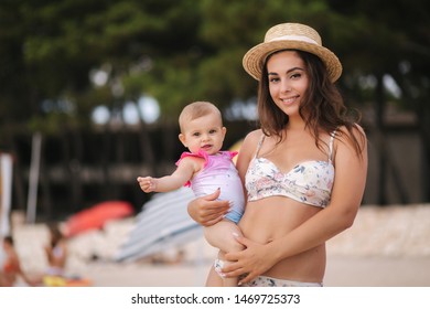 Mother Hold Baby On Hand On The Beach. Mom And Daughter In Swimsuit. Beautiful Hat