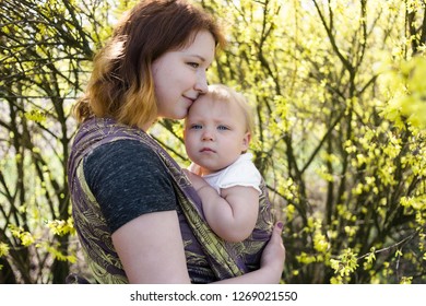 Mother Holding A Baby Girl Wrapped In A Sling, Kissing Her Forehead. High Key, Lifestyle Image Shot On A Sunny Day.