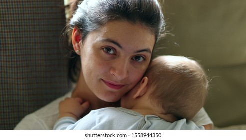 
Mother holding baby smiling to camera. Real life authentic casual mom looking to camera holding sleeping infant