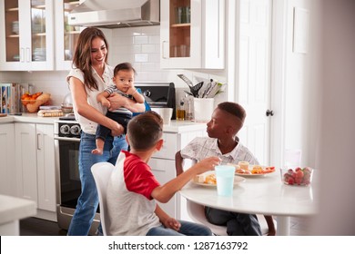 Mother Holding Baby Stands In The Kitchen Talking With Her Son And His Friend, Over For A Playdate