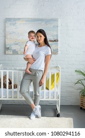 Mother Holding Cute Smiling Baby In Hands While Standing At Baby Crib At Home