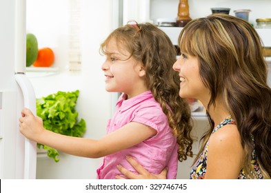 Mother Holding Up Daughter In Modern Kitchen Opening Fridge Door Looking Inside Happily.