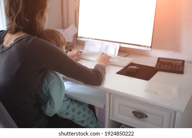 Mother holding girl kid and using computer at home with selective focus. Candid authentic and real life mom working and parenting. Home office work online