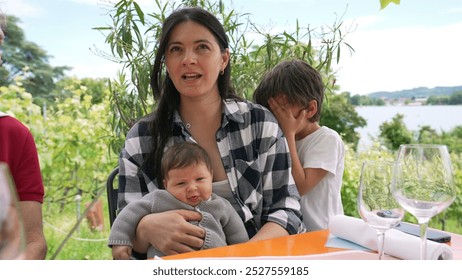 Mother holding her baby with a young child beside her, enjoying a family gathering outdoors, surrounded by greenery and a scenic lake view, a moment of warmth and togetherness - Powered by Shutterstock