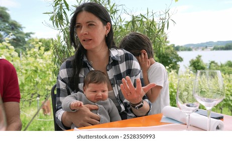 Mother holding her baby with a young child beside her, enjoying a family gathering outdoors, surrounded by greenery and a scenic lake view, a moment of warmth and togetherness - Powered by Shutterstock