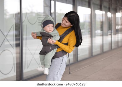 Mother holding her child in sling (baby carrier) near building outdoors - Powered by Shutterstock