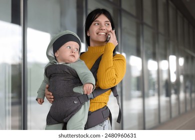 Mother holding her child in sling (baby carrier) while talking on smartphone near building outdoors - Powered by Shutterstock