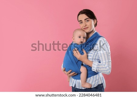 Mother holding her child in sling (baby carrier) on pink background. Space for text