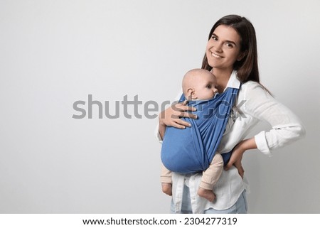 Mother holding her child in sling (baby carrier) on light grey background. Space for text