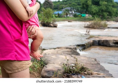 A Mother Is Holding An Infant Baby With Outdoor Waterfall Or Cataract As Blurred Background. People On Vacation And Family Time Concept Scene Photo. Selective Focus.