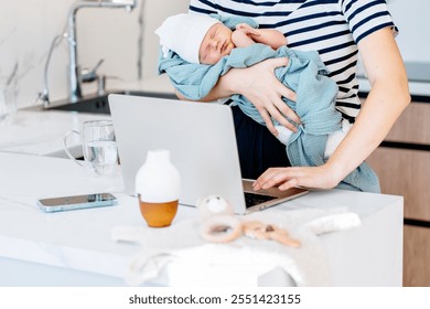 Mother holding sleeping newborn baby, working on laptop in modern kitchen. Female multitasking between motherhood and remote work, balancing career and childcare responsibilities. Parenting challenges - Powered by Shutterstock