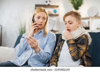 Mother Holding Thermometer And Cell Phone, Calling To Doctor, While Checking Temperature Of Her Sick Teen Son Coughing With High Fever Sitting On Sofa.