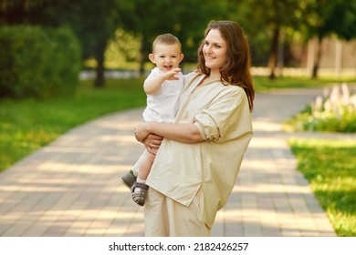 Mother Holds A Happy Toddler Boy Boy In Her Arms In The Park. Mom With A Smiling Baby In White Clothes In Nature, A One Year Old Kid