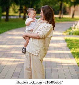 Mother Holds A Happy Toddler Boy Boy In Her Arms In The Park. Mom With A Smiling Baby In White Clothes In Nature, A One Year Old Kid