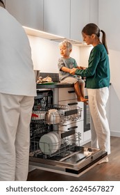 Una madre sostiene las manos de su hijo pequeño mientras el niño se sienta descalzo en el mostrador de la cocina cerca del lavavajillas abierto. El padre está cerca. Concéntrate en la vida familiar y en los momentos compartidos en casa.