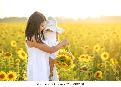 Mother Holds Little Daughter In Arms, Hiding Behind A Big Hat From The Sun In Meadow Of Sunflowers. Basket With Yellow Flowers In Mom's Hands. World Mother And Children's Day. Happy Family On Holidays