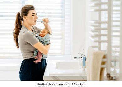 Mother holds newborn baby while brushing teeth in bright bathroom. Woman multitasks in daily routine, real-life parenting. Mother with dark brown hair tied in ponytail, wears casual home wear. - Powered by Shutterstock