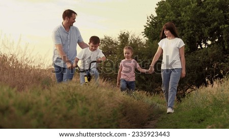 Mother holds running girl and father supports boy riding bicycle on family vacation at country area. Positive father helps boy on bicycle and mother holds active girl hand on family weekend at sunset