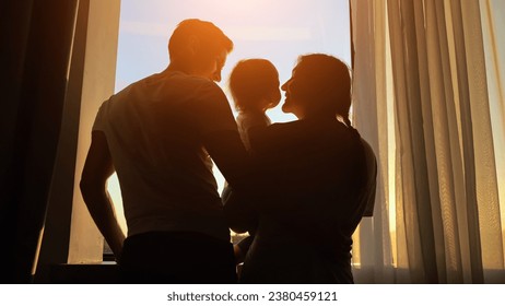 Mother holds toddler in arms near window with husband watching sunset. Family admires view mom leans down to plant kissing child cheek - Powered by Shutterstock