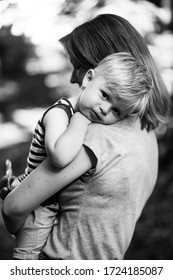 Mother Hugging Her Toddler Son And Carrying Him In The Park. Serious Little Boy Looking Straight Into The Camera With Innocent Eyes. Black And White Unposed Sincere Candid Family Love Concept.