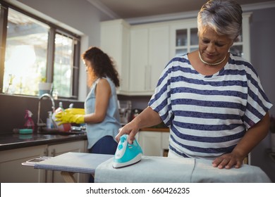 Mother ironing clothes while daughter washing utensil at home - Powered by Shutterstock