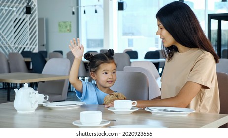 Mother With Kid Girl In Restaurant. Toddler Child With Space Buns Raises Hand With Smile Sitting By Asian Mum At Table With White Tea Cups In Local Cafe Close View