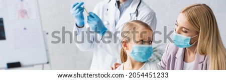 Mother and kid in medical masks near blurred family doctor with vaccine, banner