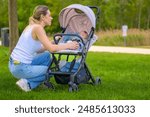 A mother kneels on the grass to adjust her baby in a stroller, enjoying a peaceful moment outdoors.