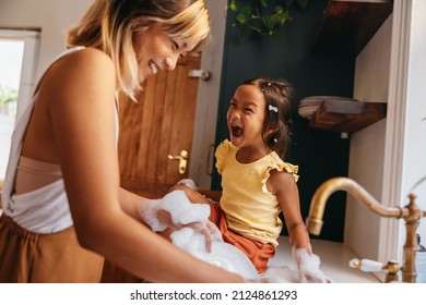 Mother Laughing With Her Daughter While Washing The Dishes In The Kitchen. Cheerful Mother And Daughter Having Fun With Soap Bubbles. Mother And Daughter Spending Quality Time At Home.