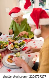 Mother In Law And Son In Law Wearing Santa Hats Sitting At Christmas Table And Eating. Family Christmas Time And Festivity Together At Home.