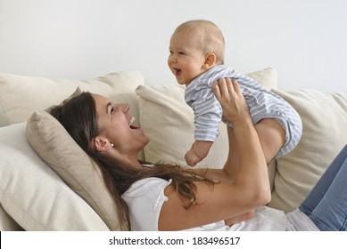 Mother Lifts Her Baby Son In The Air. She's Lying On A Sofa. They're Laughing