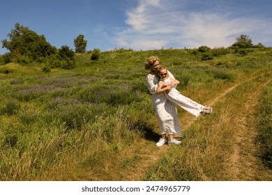 A mother in a light dress holds her little daughter under her arms and spins her around a green meadow. - Powered by Shutterstock