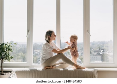 Mother and little baby sitting at home on a windowsill by the window, on a high floor. - Shutterstock ID 2148424583