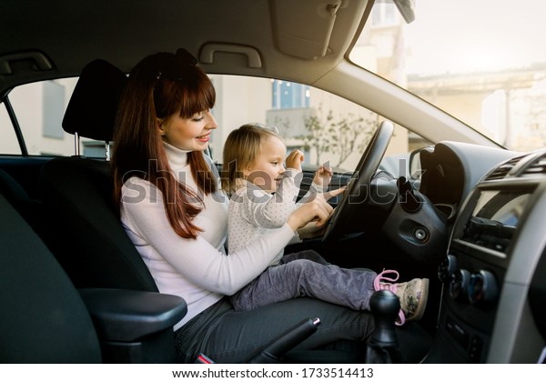 Mother with little child girl on her knees shows how\
to drive a car and what is steering wheel. Mom and baby having fun\
in a car