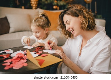Mother and little daughter making Valentine's day cards using color paper, scissors and pencil, sitting by the table in cozy room 