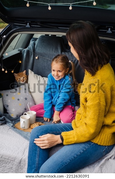 Mother with little daughter sitting in car trunk with\
a cat while traveling and eat tasty food. Family having trip with\
lovely pet