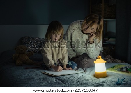 Mother and little daughter studying and drawing in a complete darkness during electricity outage. Little girl uses camping lantern to do her homework during blackout. Energy crisis concept