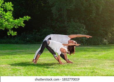 Mother and little daughter together is doing yoga asana or pilates training exercise on green grass in summer park and smiling. Concept of family outdoor sports and activity. Healthy lifestyle.
