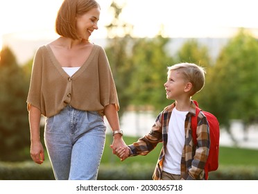 Mother And Little Son Schoolboy With Backpack Holding Hands, Going To School Together Through Park On Sunny Autumn Day, Cute Happy Boy Looking At Mom With Smile And Being Ready To Study