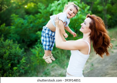 Mother With Long Curly Red Hair Playing With Her Son In The Park. Happy Family Enjoying Relaxing And Enjoying Life In Nature. Outdoor Shot. Copyspace