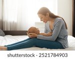 A mother lovingly gazes at her newborn while sitting in a sunlit bedroom during the afternoon