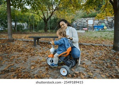 A mother lovingly guides her toddler on a tricycle through an autumn park, surrounded by fallen leaves. The warm, nurturing moment captures the essence of family bonding and outdoor play. - Powered by Shutterstock