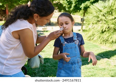 A mother lovingly wipes her daughter's face with a napkin during a sunny day at the park. The scene shows a caring family moment and a nurturing parent-child relationship outdoors. - Powered by Shutterstock