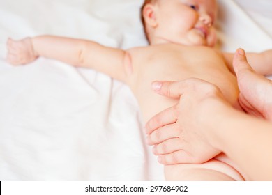 Mother Makes Massage And Stretch For Baby Stomach, With Happy Newborn At White Bed, Apply Oil On The Hand