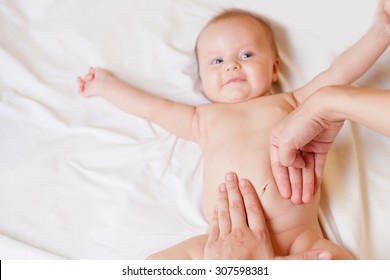 Mother Makes Massage And Stretch For Baby Stomach, With Happy Newborn At White Bed, Apply Oil On The Hand