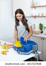 Mother Making Breakfast For Her Children In The Morning And A Snack For School. She Puts Yellow Plastic Lunch Box To Her Son In A School Backpack.