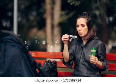 Mother Making Soap Bubbles To Entertain Baby In Stroller. Young Cool Mom Having Fun With Newborn Outdoors
