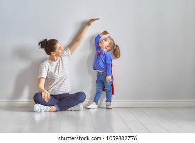 Mother is measuring growth of child daughter near empty wall. Girl in superhero costume.